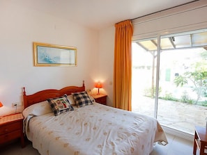 A bedroom with a bed, a bedside table with a lamp, a window with curtains, and a painting on the wall.