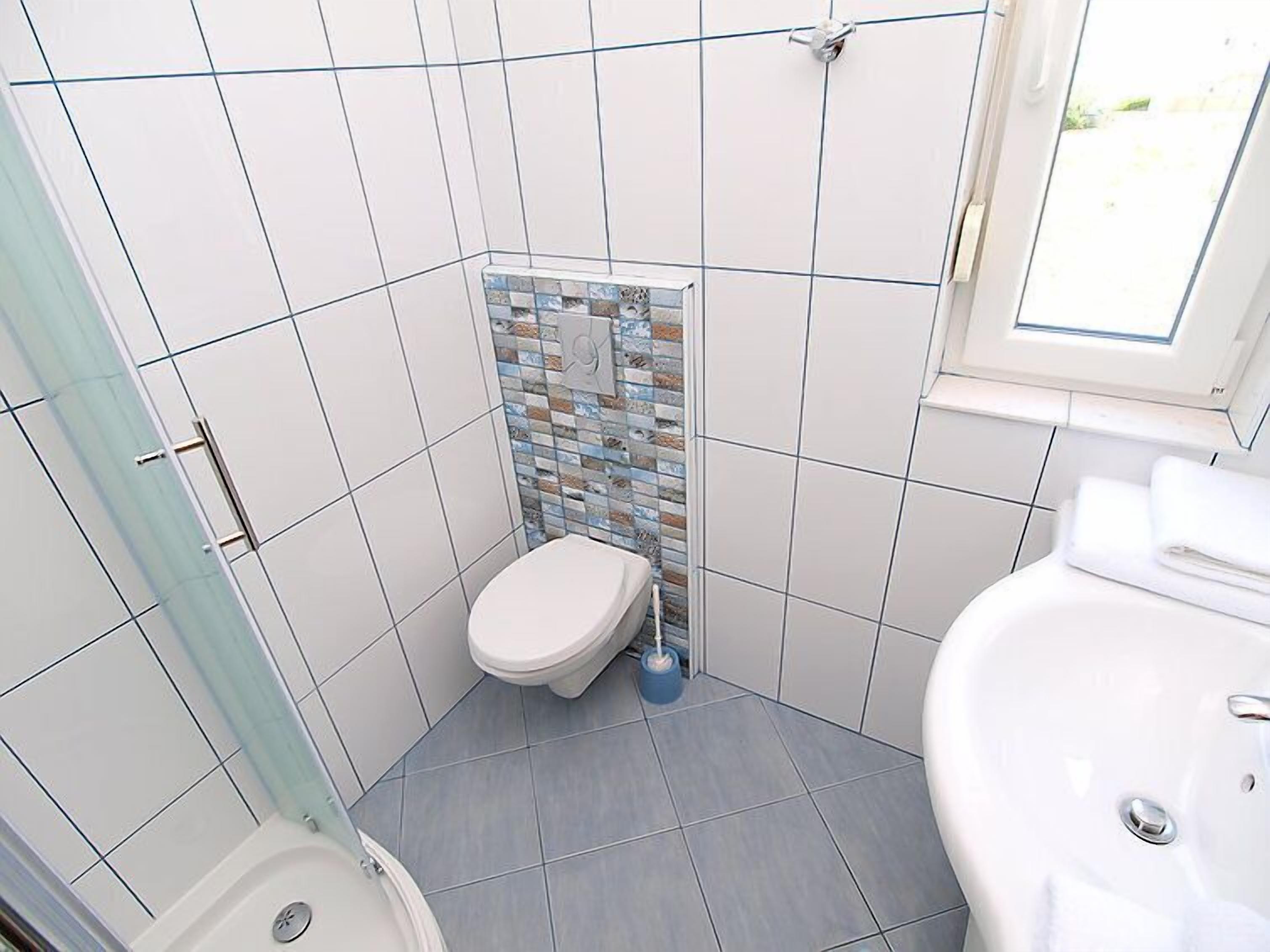 A small bathroom with a toilet, a sink, and a shower. The walls are tiled, and there is a window above the sink.