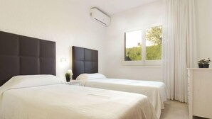 Habitación de hotel con dos camas, una mesita de noche, un ventanal con cortinas y un pequeño escritorio con planta en maceta.