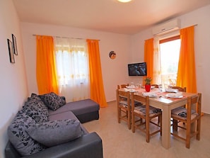 A modern living room with a grey sofa, orange curtains, a dining table set for four, and a flat-screen TV on the wall.