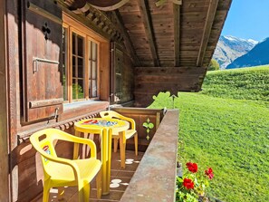 Een houten chalet met een balkon, twee gele plastic stoelen, een kleine ronde tafel en uitzicht op een groene heuvel en bergen.