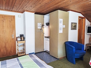 Een kleine kamer met een kitchenette, een blauwe stoel en een houten plafond.