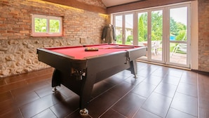 Une table de billard dans une pièce aux murs en briques, aux grandes fenêtres et qui offre une vue sur la verdure à l’extérieur.