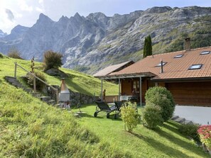 Uma cabana na montanha com telhado inclinado, chaminé e um pequeno terraço com móveis de exterior.