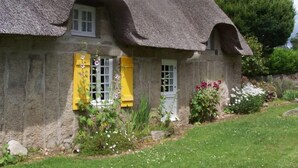 Ein Steinhäuschen mit einem Strohdach, zwei Fenstern mit Fensterläden und einem Garten mit Blumen und Sträuchern.