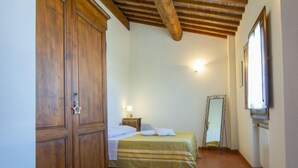 A bedroom with a wooden wardrobe, a bed with striped bedding, a bedside table with a lamp, a mirror, and a window with blinds. - Borgo Fontana by Interhome (Vinci)