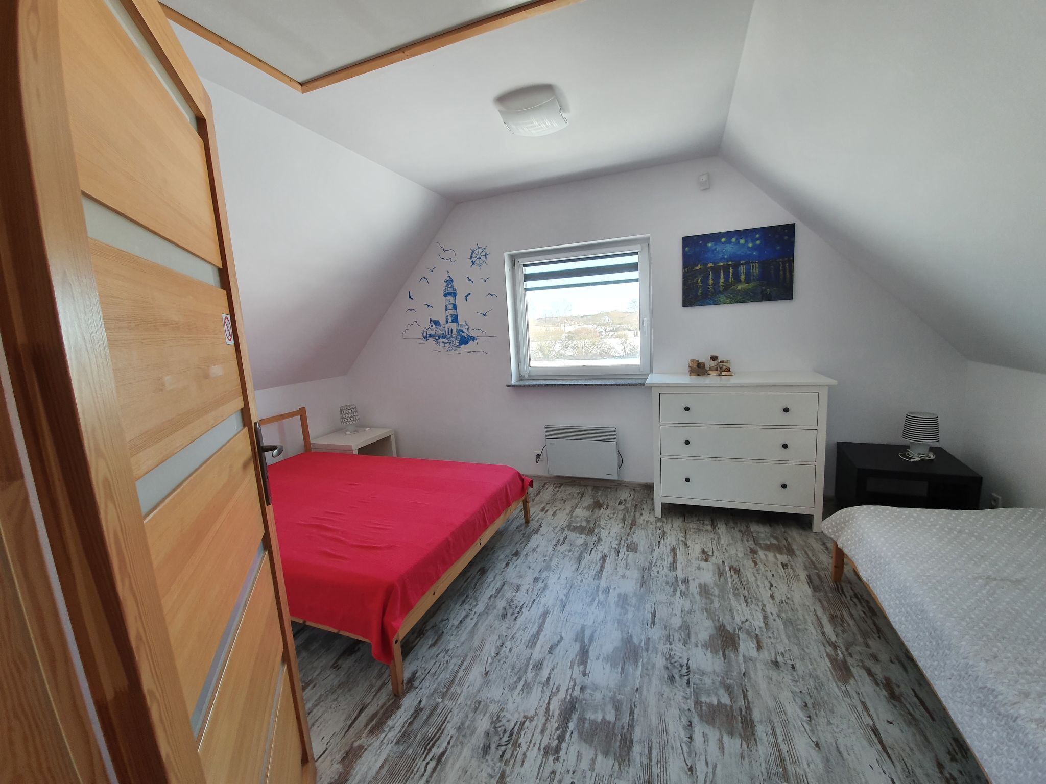 A small, well-lit room with a sloped ceiling, a bed with a red bedspread, a white dresser, a small table with a lamp, and a window with a view of the outside.