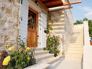 Steps lead to a stone-clad entrance with a wooden door, surrounded by vibrant flowering plants.