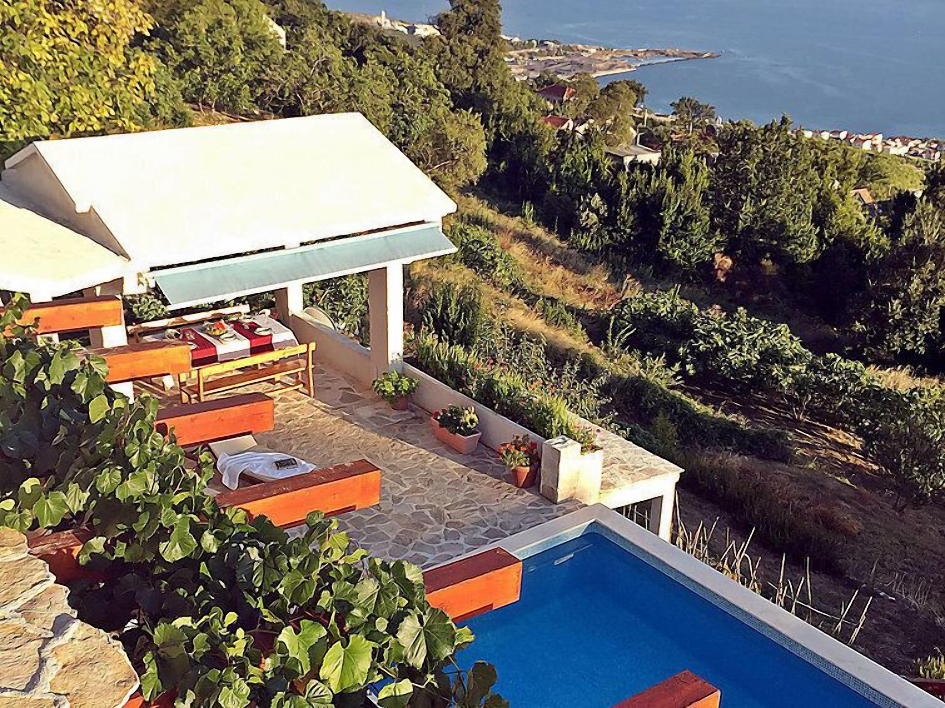 A poolside area with a covered seating area, stone flooring, and a view of the sea.