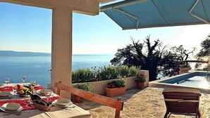An outdoor dining area with a table set for two, overlooking a pool and a scenic view of the sea.