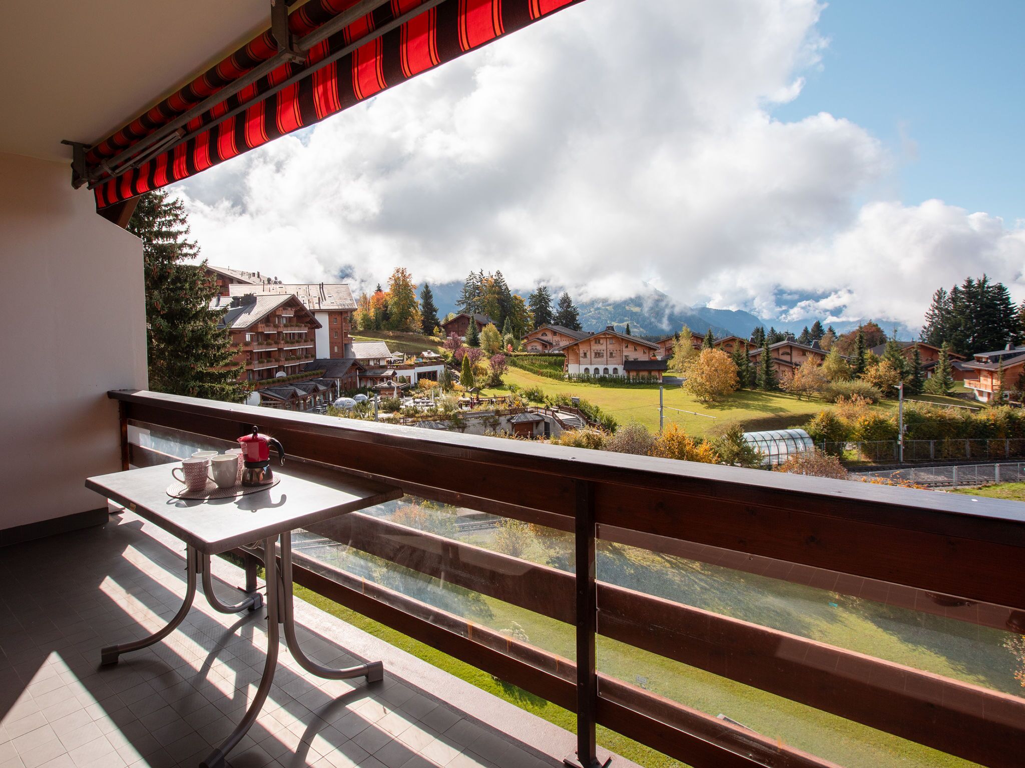 A balcony with a table and chairs, overlooking a mountainous landscape with buildings and a clear sky.