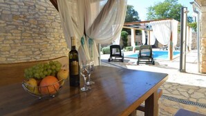 A wooden table with a bottle of wine, two glasses, and a bowl of fruit. There are two chairs and a pool in the background. - Vacation home Marija by Interhome (Oklaj)