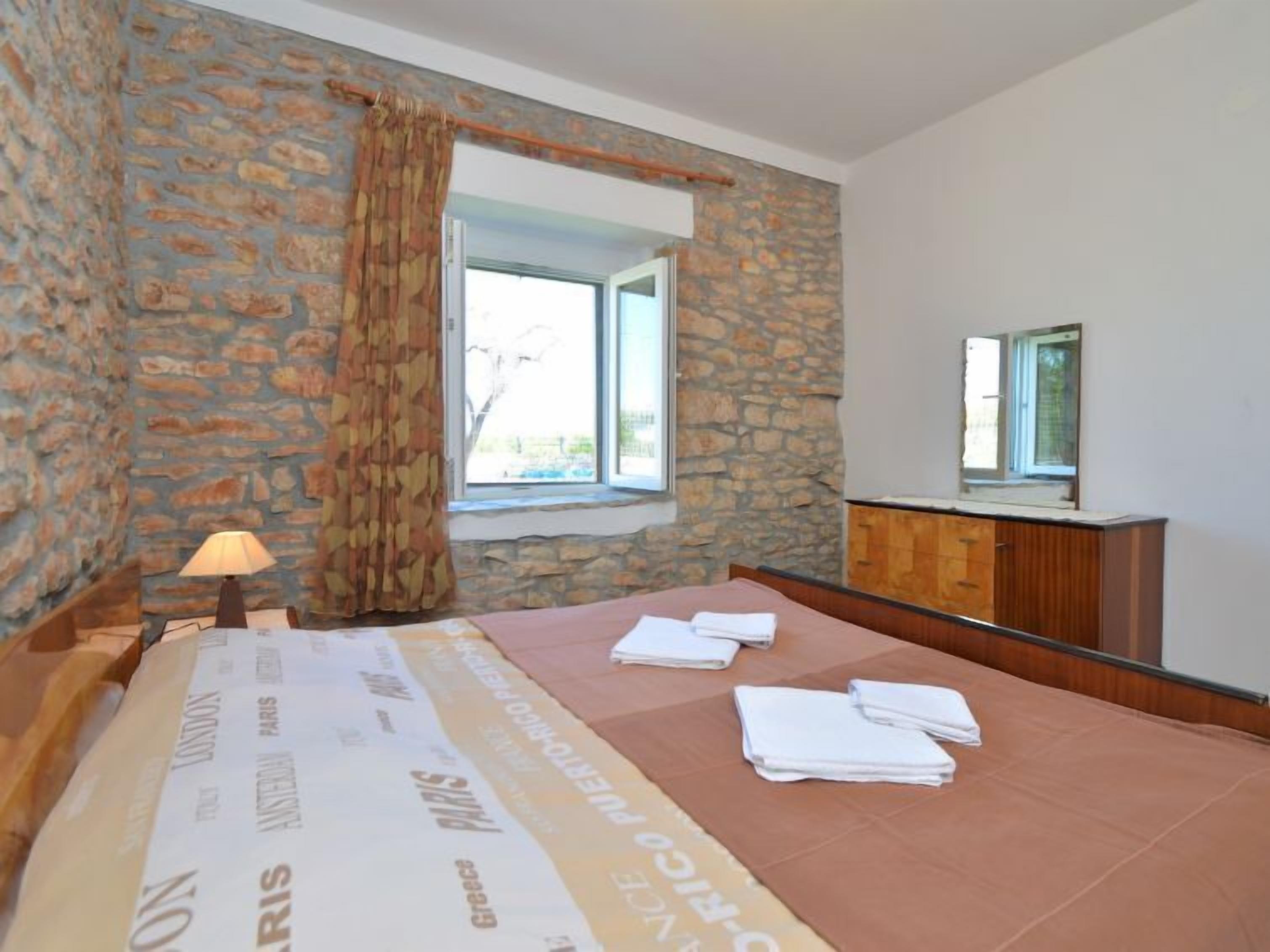 Un dormitorio con paredes de piedra, una ventana con cortinas, una cama con manta, una lámpara de noche y un armario de madera.