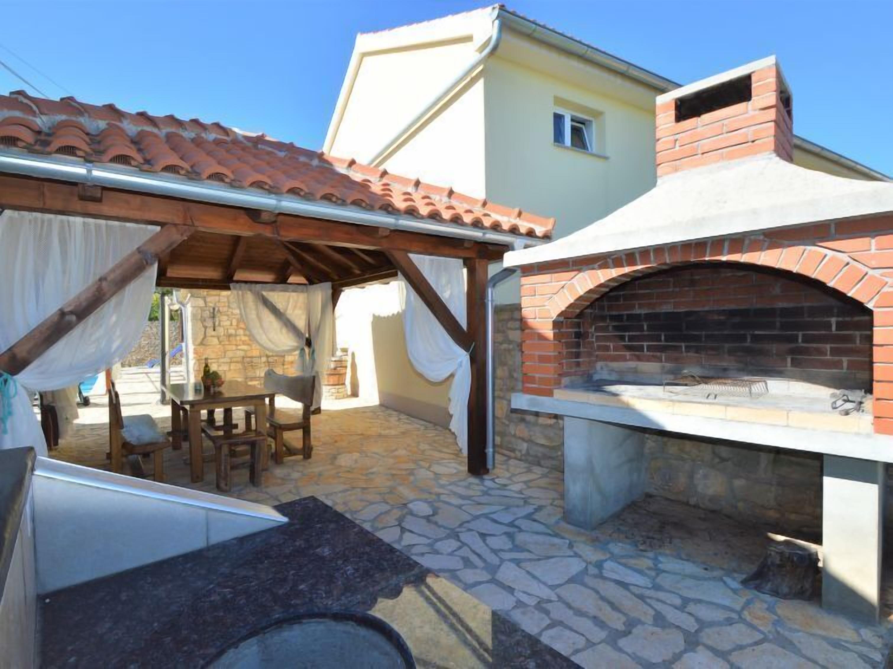 Une terrasse extérieure avec un sol en pierre, un four en briques, une table avec des chaises et un espace salon abrité.
