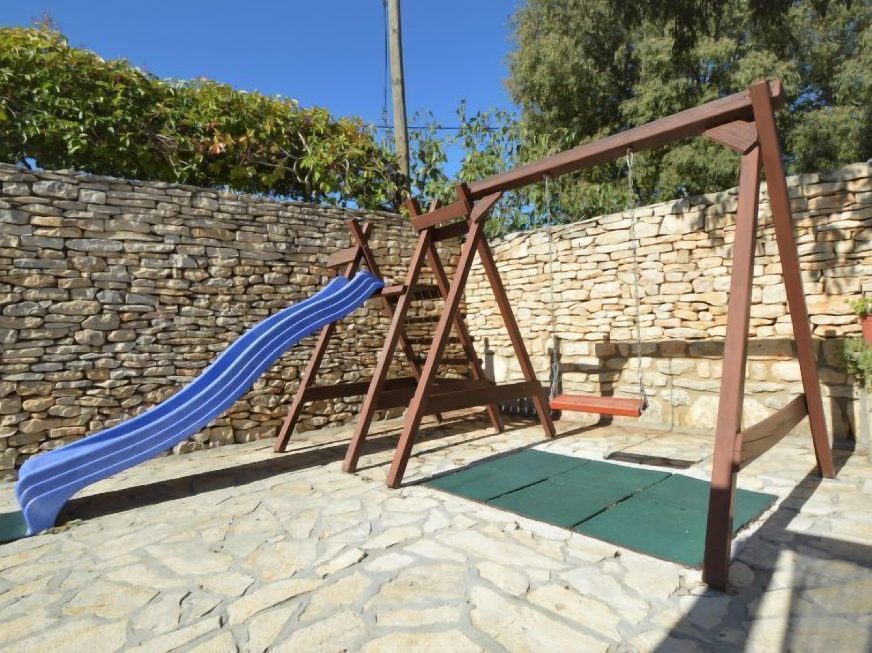 A wooden swing set with a blue slide in a stone-walled outdoor area.
