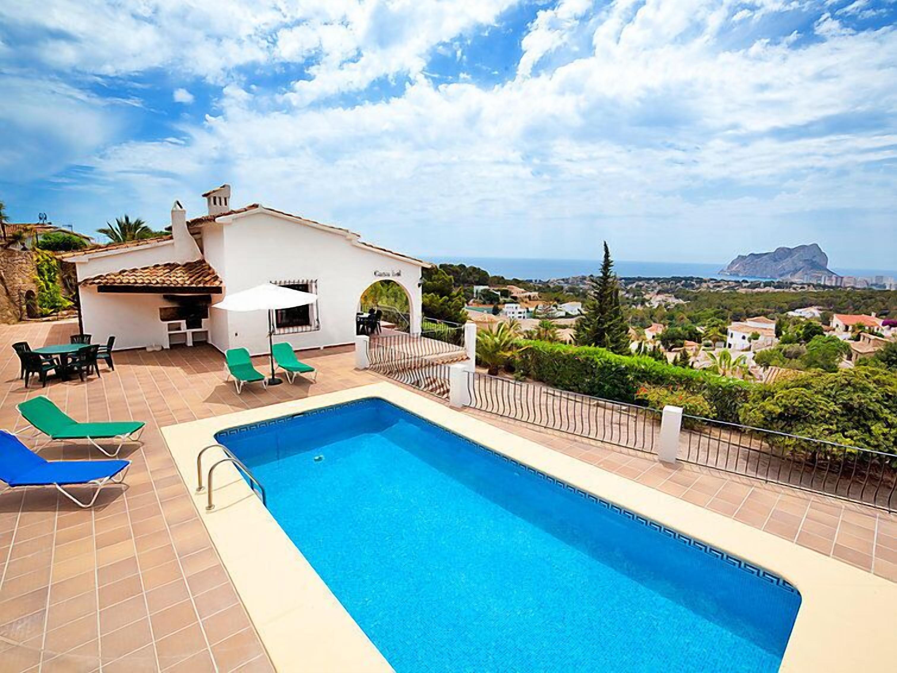 A swimming pool with lounge chairs, a barbecue area, and a view of the sea and town.