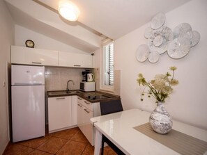 A compact kitchen with white cabinets, a built-in oven, a microwave, and a coffee maker. There's a dining table with a vase of flowers and a window with blinds.