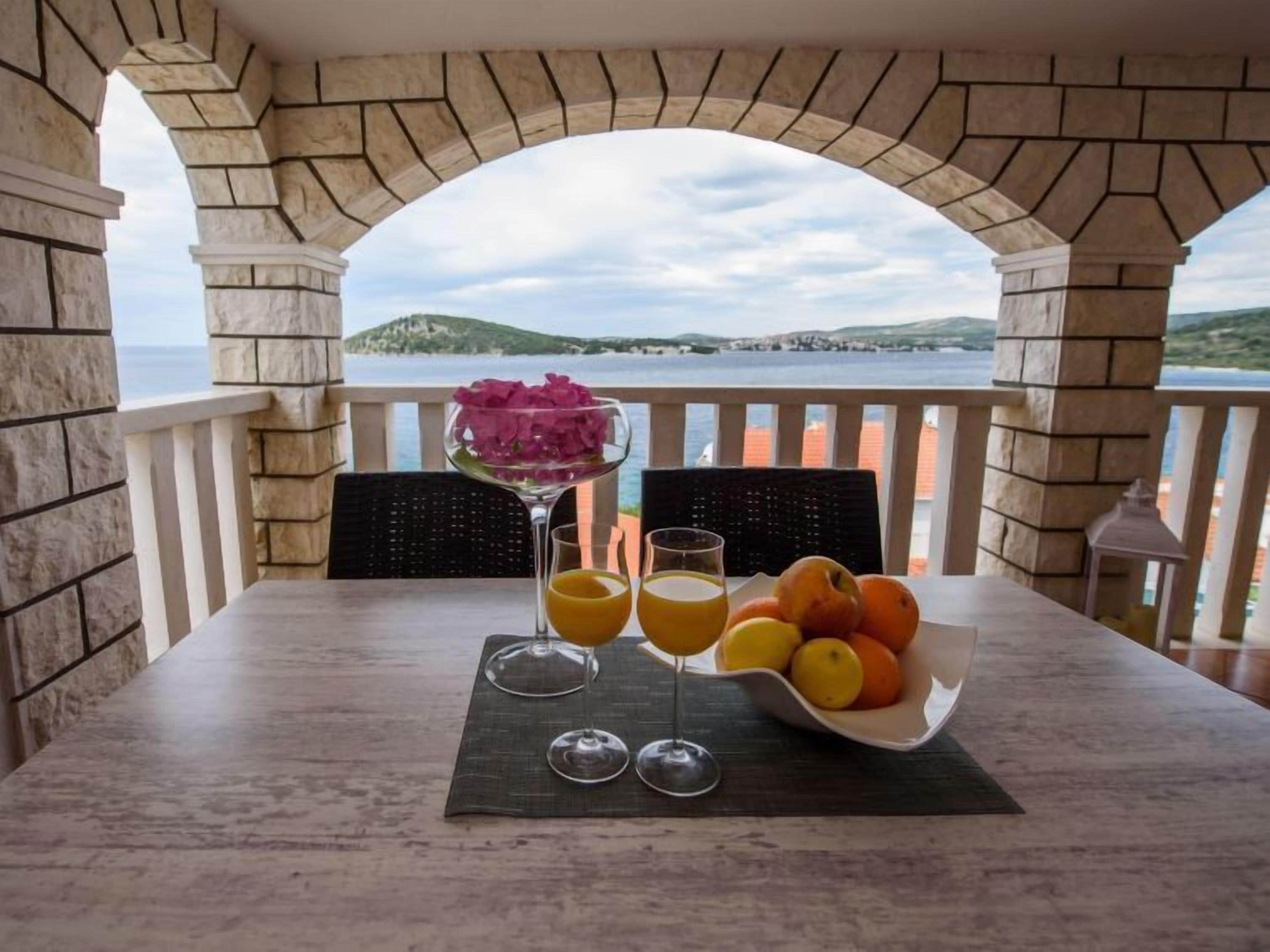 Ett bord med en fruktskål, två glas apelsinjuice och ett martiniglas med rosa blommor, placerat med en vacker utsikt över havet och kullarna i bakgrunden.
