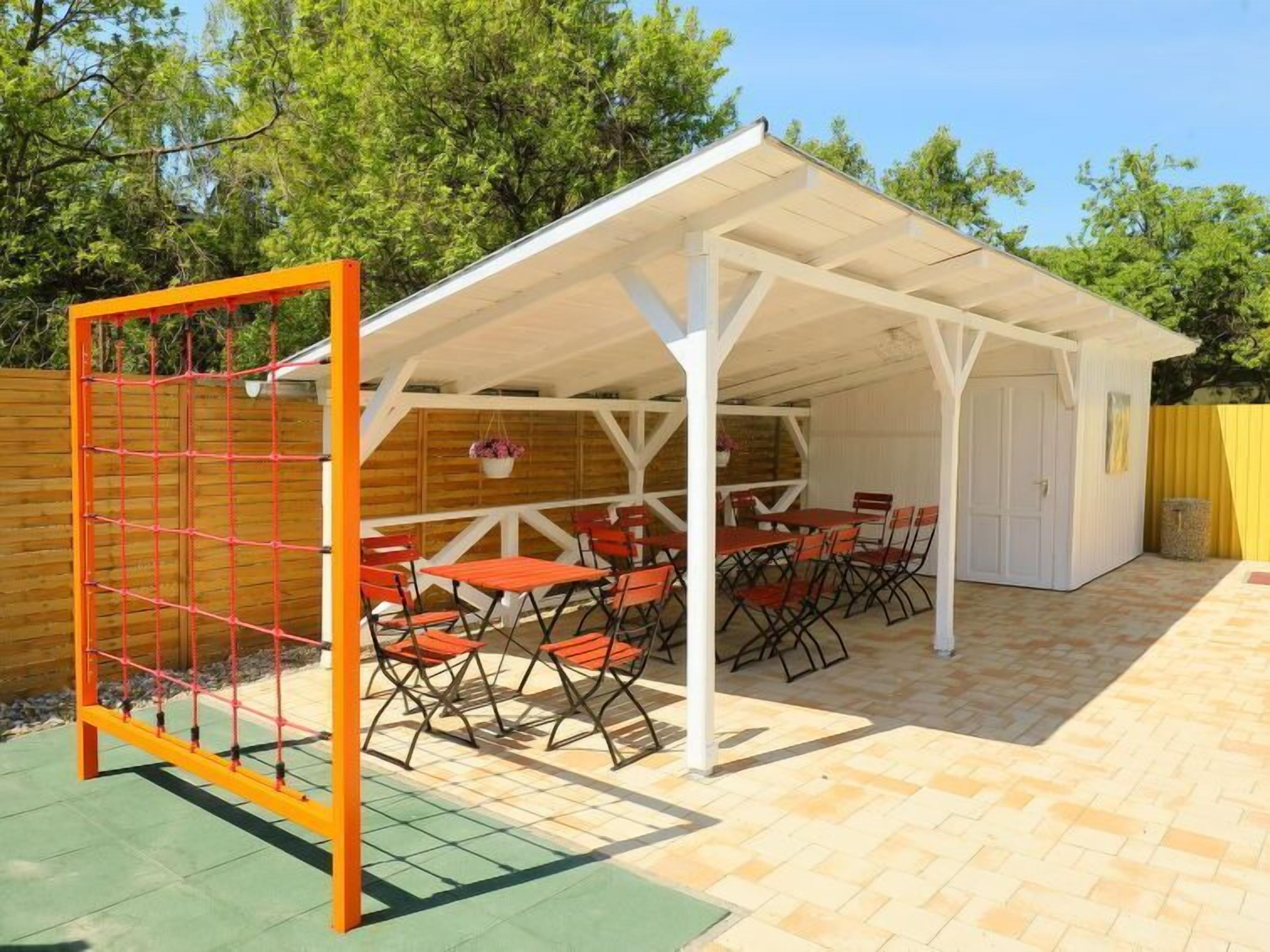 A covered patio area with tables and chairs, a wooden fence, and a white building in the background.