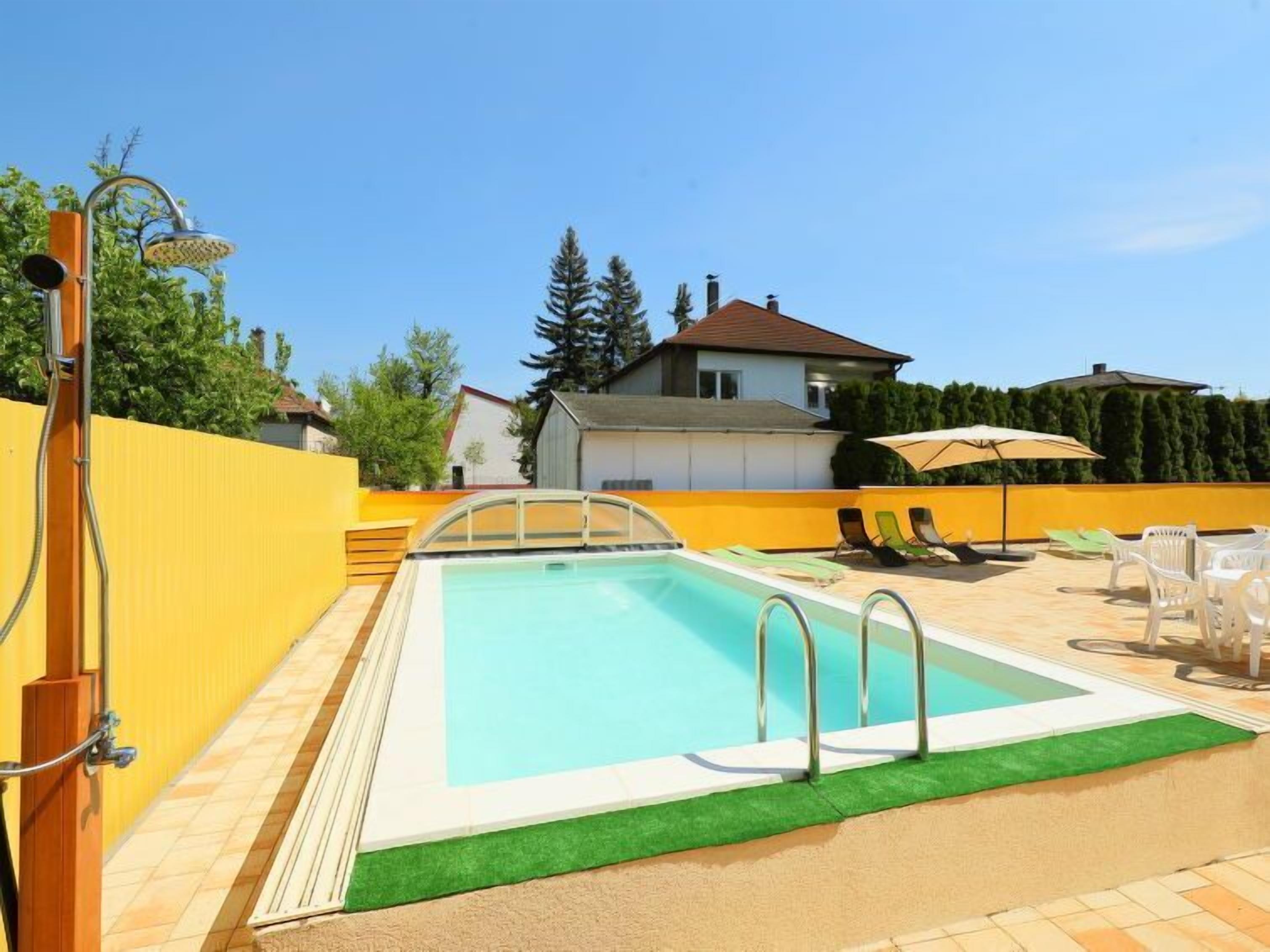 Una piscina rectangular con borde verde y zona de sillones de baño con sillas y una sombrilla.