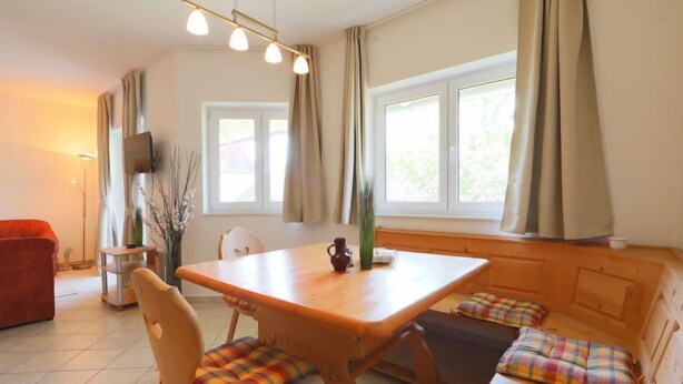 A cozy room with a wooden dining table, two chairs, a bench with colorful cushions, a red sofa, and a TV on the wall.