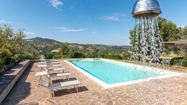 Ein Swimmingpool mit einer Steinterrasse und Liegestühlen, umgeben von Grünflächen und Hügeln.