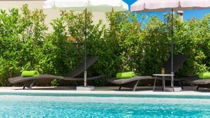 Zona de piscina con sillones de descanso y sombrillas, rodeada de vegetación.