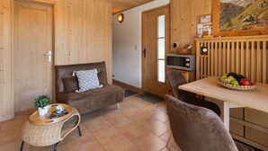 Ett mysigt rum med träväggar, en brun soffa, ett litet runt bord med en växt, en flätad stol och ett matbord med en fruktskål.