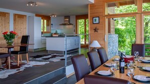 Una cucina moderna con isola centrale, un'area pranzo con un tavolo lungo e sedie, e vista su una vegetazione rigogliosa attraverso ampie finestre.