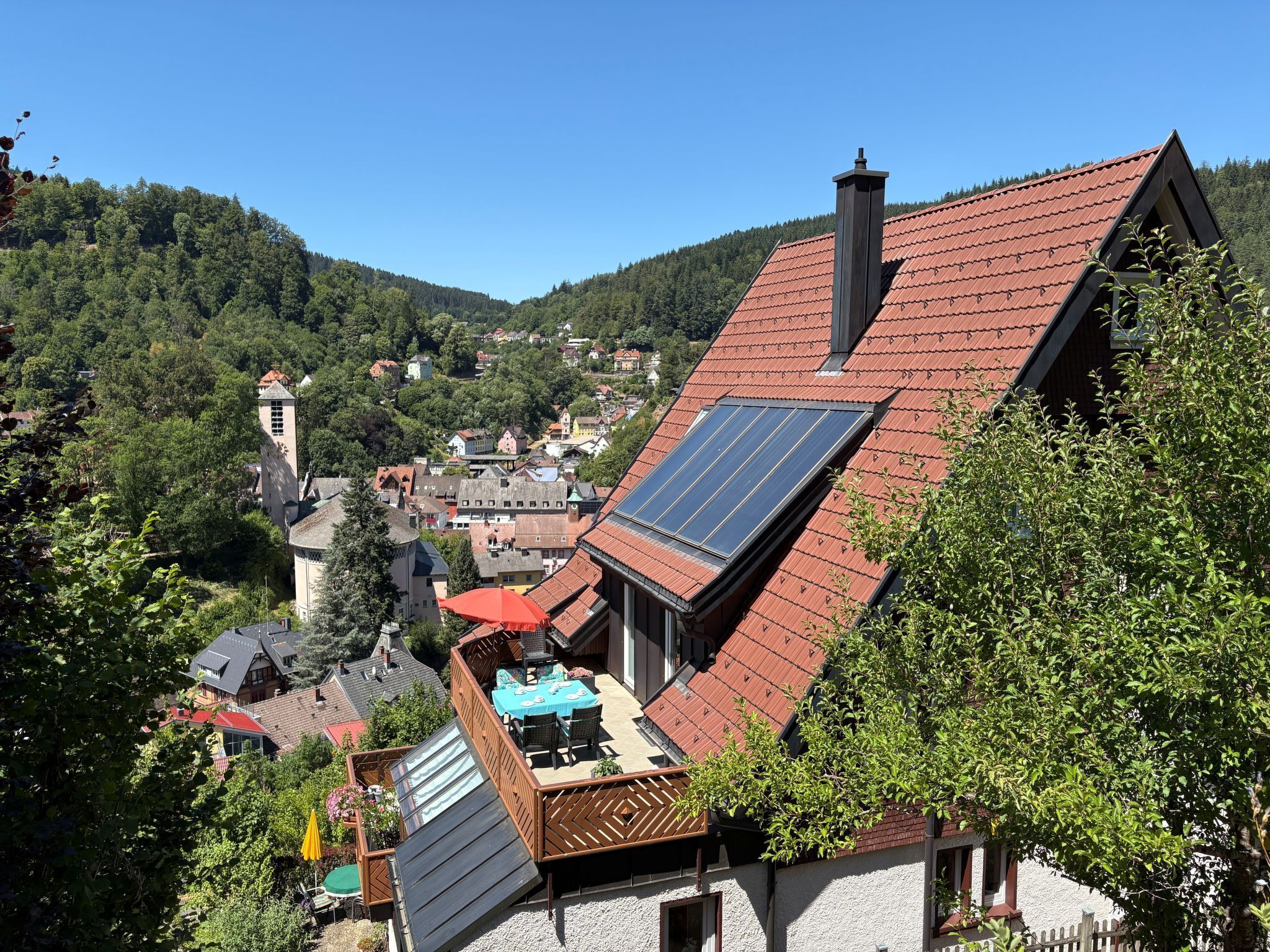 A residential building with a sloped red roof, solar panels, and a visible balcony with outdoor furniture.