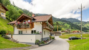 En tvåvånings chalet med ett sluttande tak, träbalkonger med blomlådor och en asfalterad uppfart som leder till en bergsväg.