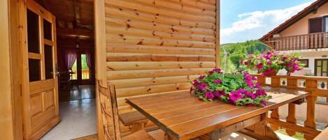A wooden log cabin with a wooden table and chairs, a balcony with flowers, and a view of greenery.