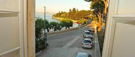Une rue côtière bordée de voitures garées, un ciel dégagé et une vue sur la mer et les montagnes lointaines.