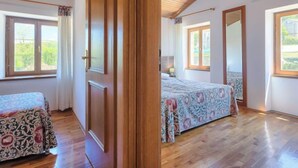 Ein Schlafzimmer mit Holzboden, zwei Betten mit gemusterten Bettdecken, einem großen Spiegel und Fenstern mit Blick auf Grünflächen.