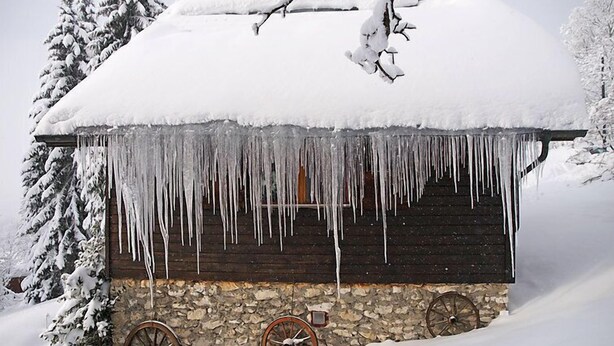 Une cabane en bois, avec des glaçons pendants du toit, entourée d’arbres couverts de neige.