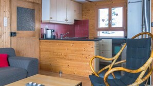 Une pièce chaleureuse avec un intérieur en bois, un canapé gris, un fauteuil en osier, une petite table et un coin cuisine doté de meubles en bois et d’un plan de travail rouge.
