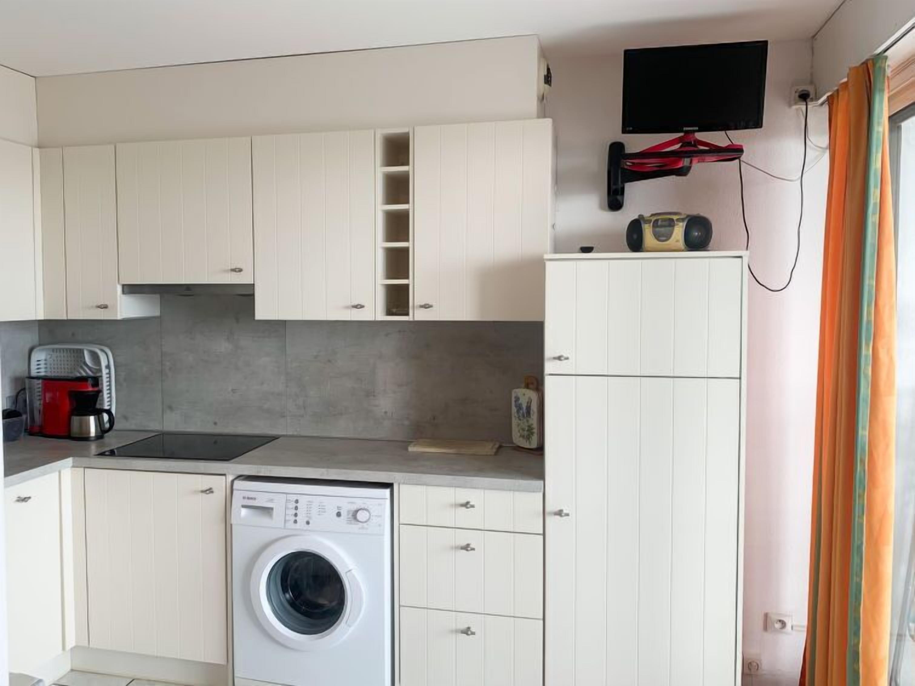 A compact kitchen with a washing machine, a coffee maker, and a flat-screen TV mounted on the wall.