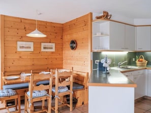 Un intérieur en bois comprenant une table à manger, des chaises, un luminaire suspendu, un coin cuisine avec un évier et un plan de travail, ainsi que des tableaux encadrés accrochés au mur.