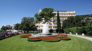Une place publique bien entretenue, avec une fontaine centrale, entourée de haies soigneusement taillées et d’une variété d’arbres. Au fond, on aperçoit un immeuble à plusieurs étages doté de balcons.