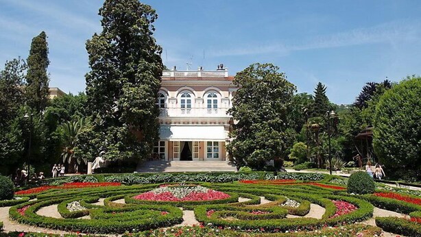 Un grand bâtiment historique à la façade symétrique, doté d’un jardin à la française avec des parterres de fleurs aux formes géométriques.
