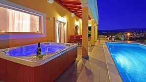 Un espace piscine comprenant un jacuzzi et une piscine, un patio couvert et une vue sur la ville la nuit.