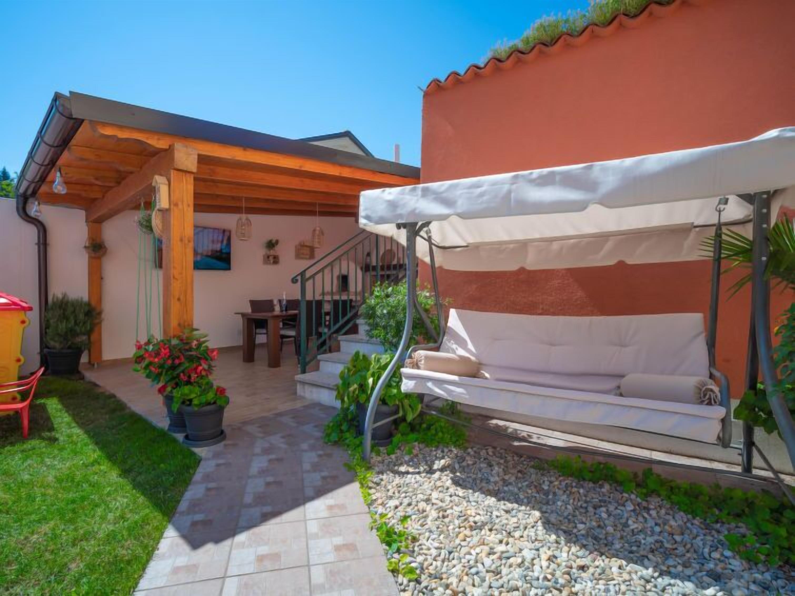 A covered outdoor seating area with a cushioned bench, a staircase leading to an upper level, and a patio with potted plants and a tiled walkway.