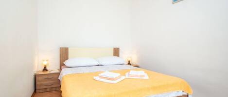 Una cama bien hecha con una colcha amarilla, almohadas blancas y un cabecero de madera. Hay dos mesitas de noche con lámparas, y un cuadro enmarcado en la pared.