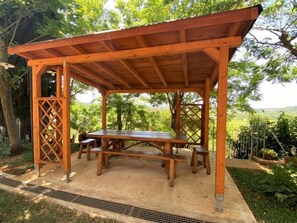 Une pergola en bois avec une table de pique-nique et des bancs, entourée de verdure et d’arbres.