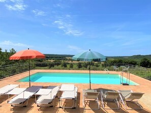 Une piscine entourée d’une clôture et de verdure, avec des chaises longues et des parasols.
