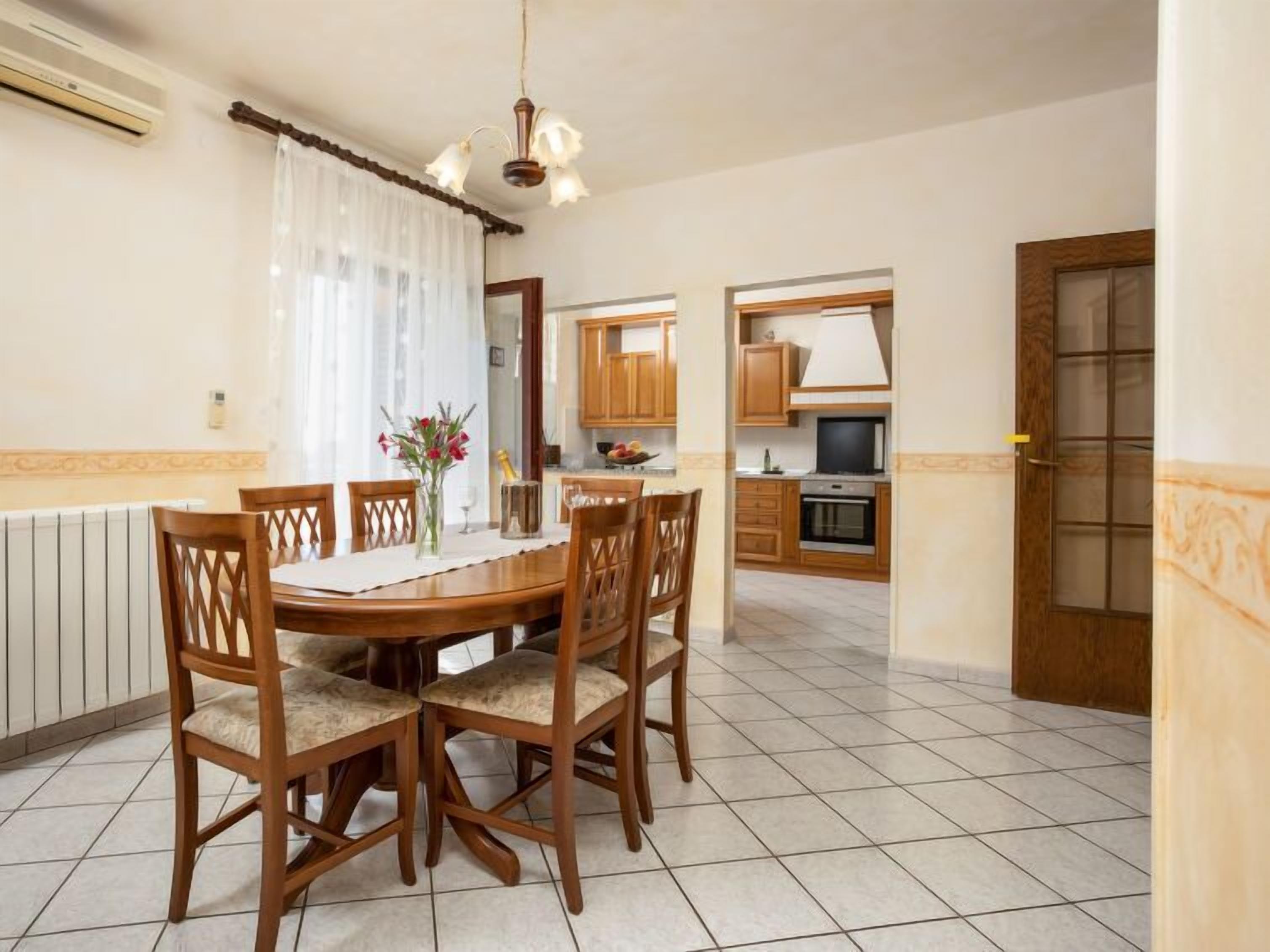 Un comedor con una mesa y sillas de madera, una araña y una cocina al fondo.
