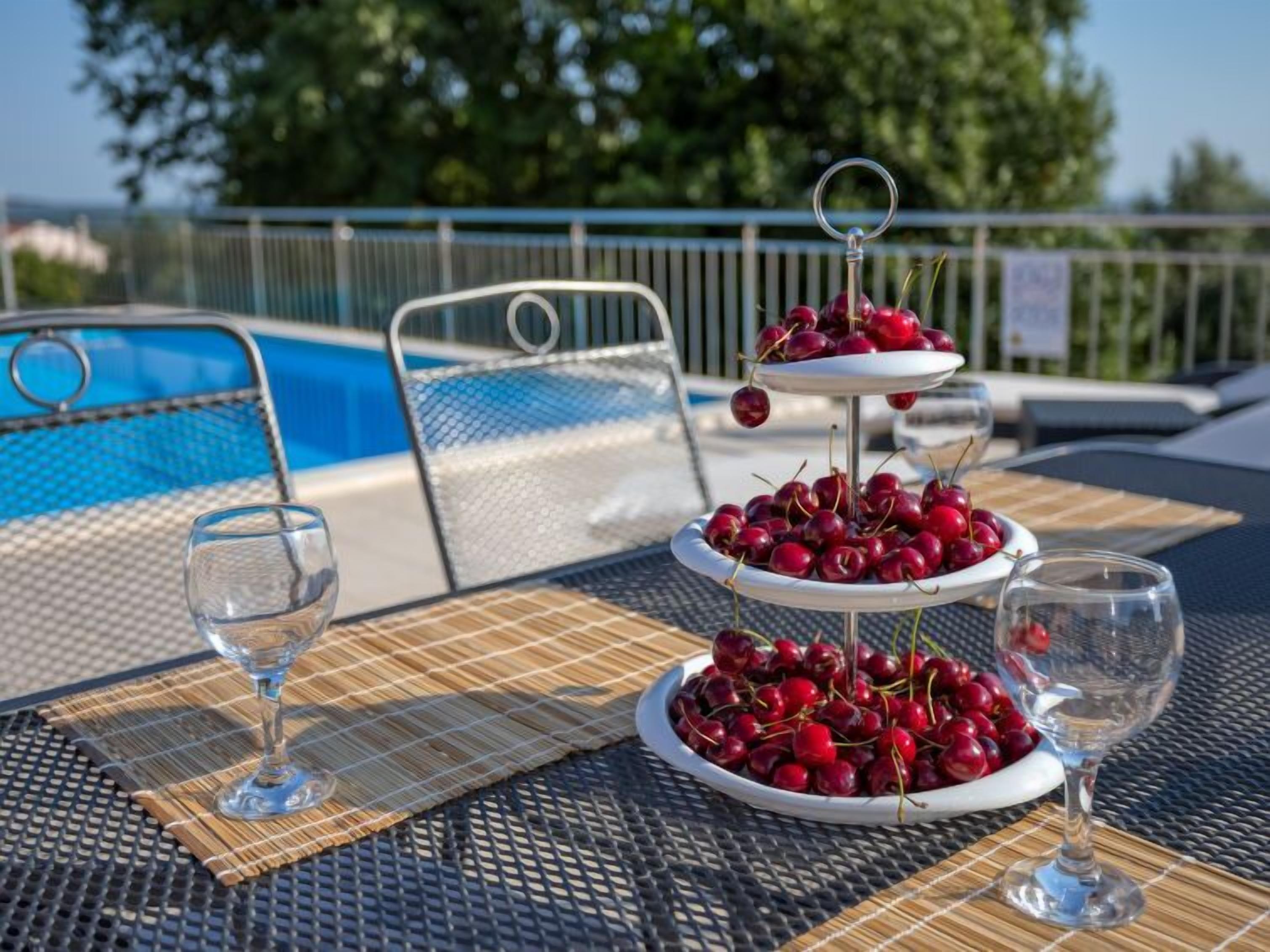 Et bord ved poolen med et frugtfad i tre etager, to glas og en pool i baggrunden.