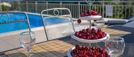 A poolside table with a three-tiered fruit stand, two glasses, and a pool in the background.