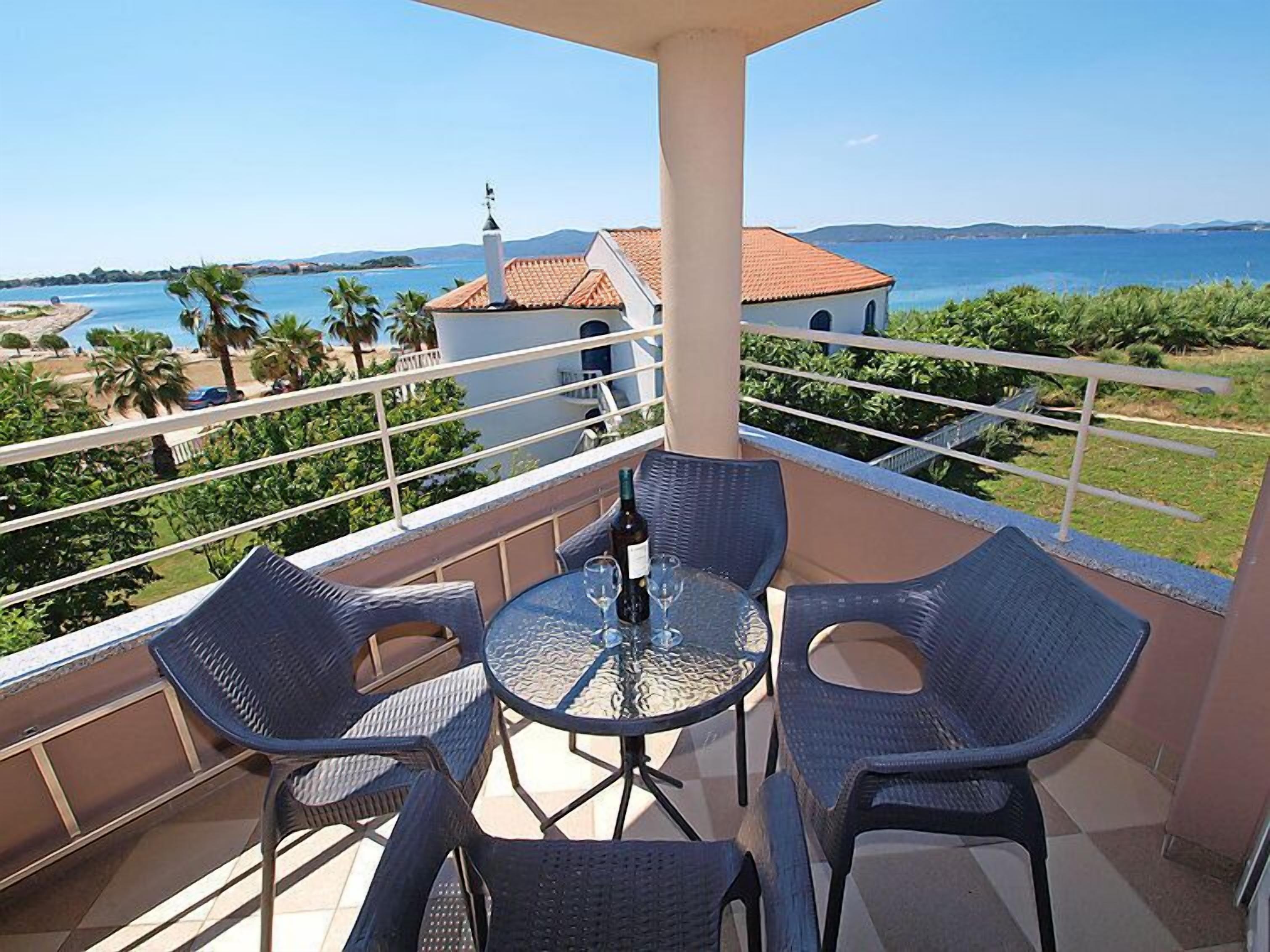 A balcony with wicker chairs and a glass table, overlooking a coastal view with a building and palm trees.