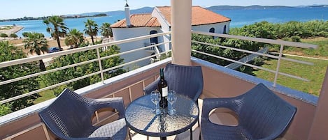 A balcony with wicker chairs and a glass table, overlooking a coastal view with a building and palm trees.